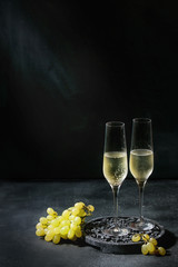 Two glasses of white champagne served on black wooden decorative board with green grapes over dark texture background. Copy space