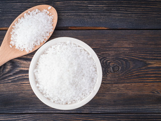 White bowl with large sea salt and wooden spoon on brown dark  burnt wooden table. Top view.