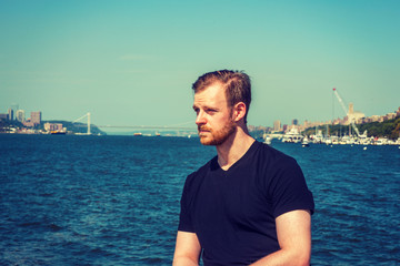 Fototapeta premium American man traveling in New York. Wearing black v neck T shirt, a young guy with beard, mustache standing by Hudson River, looking forward. Bridge, harbor on background. Filtered effect..