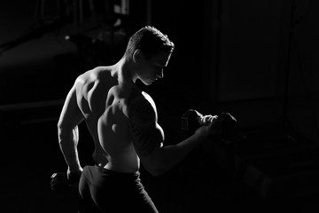 Handsome power athletic guy bodybuilder doing exercises with dumbbell. Fitness muscular body on dark background.