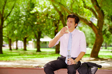 Handsome young businessman wearing a purple t-shirt and eating a sandwich and holding a bottle of water with his other hand at outdoors, in a blurred park background