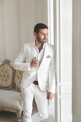 Elegant young handsome man with a beard in a white classic suit. The hotel's interior. Photo...
