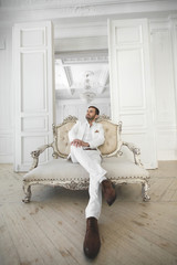 Elegant young handsome man with a beard in a white classic suit. The hotel's interior. Photo...