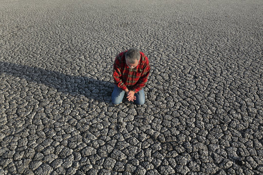 Desperate man kneeling and praying at dry cracked land after drought, natural disaster