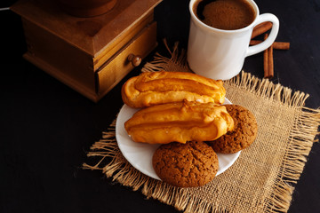 A cup of coffee, eclairs and oatmeal cookies on a black textured background.