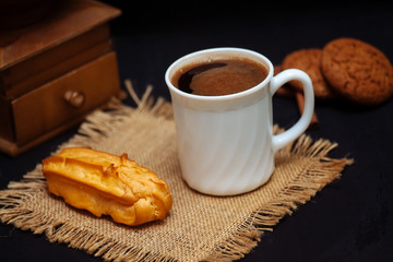 A cup of coffee, eclairs and oatmeal cookies on a black textured background.