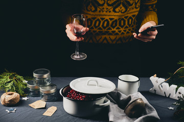 Woman hands with glass of wine and smart phone in front of Christmas themed table with ingredients for cranberry jam