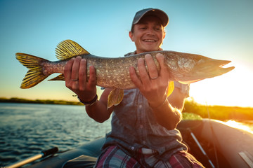 Happy angler holds pike fish sitting in a boat with river on the background