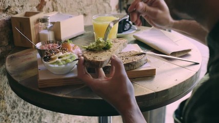 Hipster millenial man enjoys delicious and healthy breakfast at downtown brunch spot or coffee shop on lazy sunday afternoon. Eats toast with avocado spread, and drinks fresh orange juice