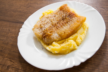 Fried fish fillet of cod with mashed potato in white plate on wooden background.