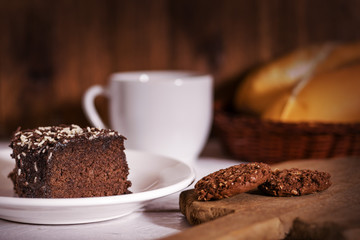 Chocolate cake, cookies, coffee porcelain cup and bread loafs on top of a wooden white table top and wood background