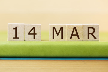 Wooden cube shape calendar for MAR 14 on green book, table.