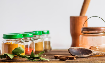 Spices and seasoning containers, glass pots and bowls in a row, on a wooden surface, with mortar and pestle, bay leaf, wooden spoon and cinnamon decorating, white background