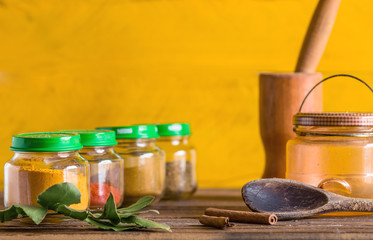 Spices and seasoning containers, glass pots and bowls in a row, on a wooden surface, with mortar and pestle, bay leaf, wooden spoon and cinnamon decorating, yellow background