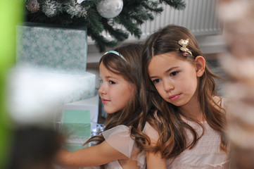 Two girls on sofa next to a Christmas tree talking and play on mobile phones. Sisters in living room waiting a New Year