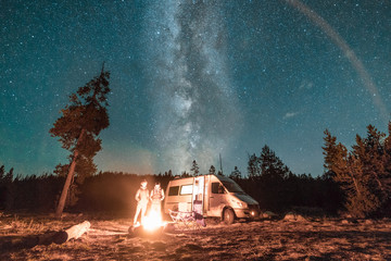 Vanlife couple standing in front of campfire and van under Milky Way night sky.  © Matt