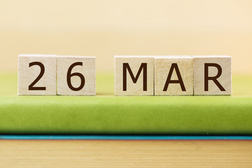 Wooden cube shape calendar for MAR 26 on green book, table. 