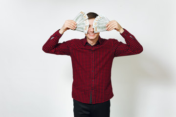 Naklejka premium Handsome cheerful positive satisfied caucasian young happy business man 25-30 years in red shirt hiding behind wad of cash money on white background isolated for advertisement. Winner with dollars.
