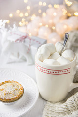 White Mug with Hot Chocolate Cocoa Drink and Marshmallows on Top Mince Pie on Plate. Sparkling Garland Lights in Background. Knitted Sweater Gift Box Christmas New Year Advent Morning Cozy