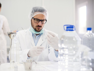 Close up of serious focused professional scientist bearded young man working at laboratory desk with bottles of water and flasks in front.