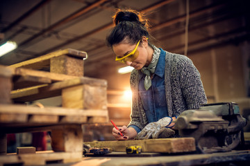 Portrait view of satisfied smiling middle aged professional female carpentry worker with a pencil drawing on the wood and tape measure, steel vise on the table in the workshop.