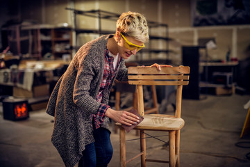Attractive satisfied female furniture designer carefully sanding a chair frame with shelves of...