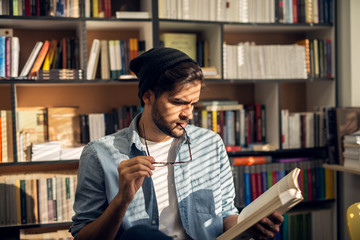 Portrait view of young curious bearded hipster student guy reading book in the library with...