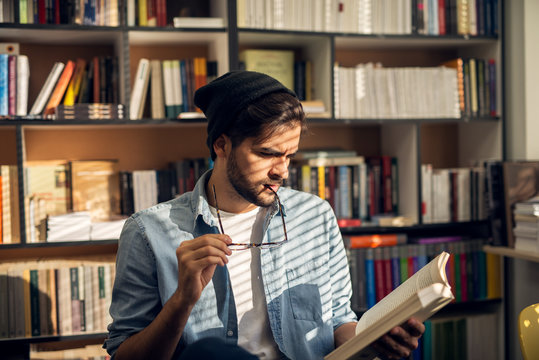Portrait view of young curious bearded hipster student guy reading book in the library with eyeglasses in a mouth near the window.