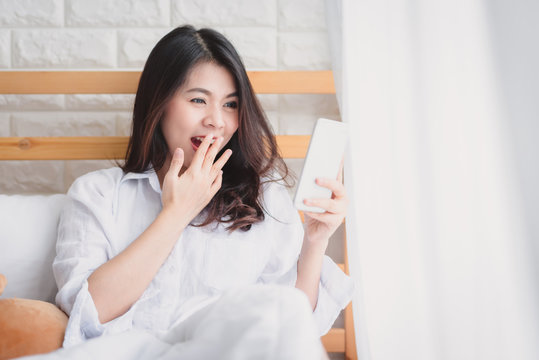 Beautiful Asian woman surprised and shocked while looking in smartphone mobile on the bed in the morning after wake up