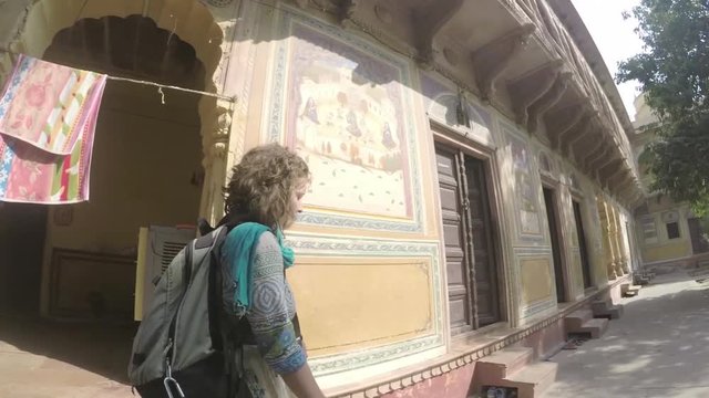 Caucasian woman walks through grounds of beautiful temple to Goddess Radha in India, smooth tracking shot, with traditional artwork on walls. Wide angle. 2X slow motion.
