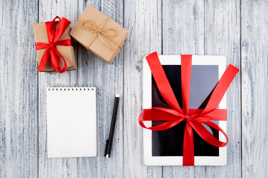 gifts and a tablet wrapped in a red ribbon, a paper notepad, a pen on a white vintage wooden background