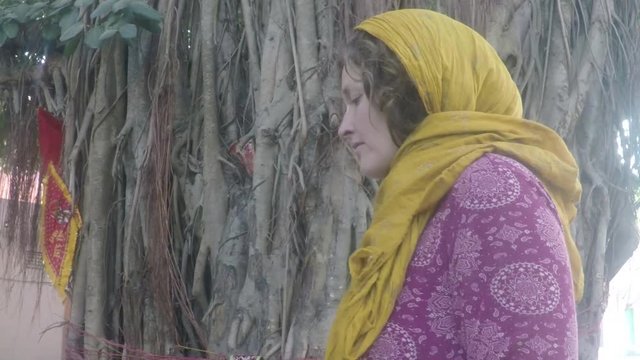 ]Caucasian woman pilgrim walks around a sacred banyan tree with temple in background, smooth tracking shot, four times slow motion, in India.