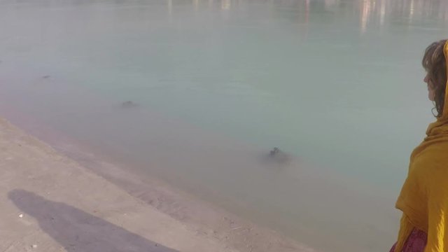]Woman pilgrim, caucasion, walks the banks of the Ganges River in Rishikesh, back side view, looking thoughtful. Smooth tracking shot, four times slow motion.