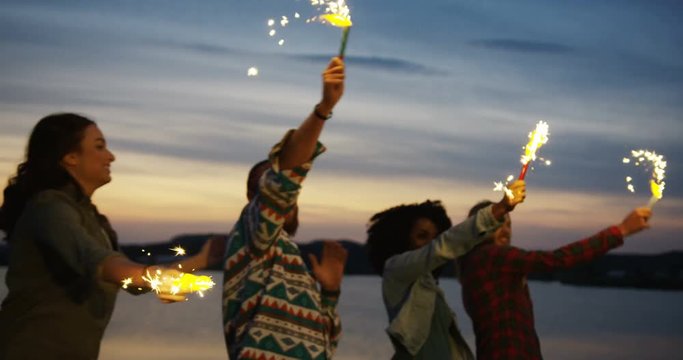 Young multi ethnical hipsters having fun and swaying sparkler sticks in the twilights near the big lake. Outside
