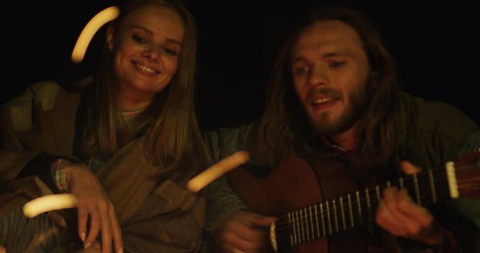 Close up of the loving couple warming near the fire. Blonde beautiful woman listening to the man with a long hair playing the guitar. At night. Outdoor