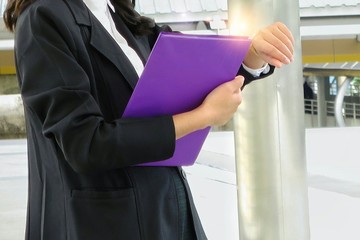 Obraz premium A young business woman in black suit looking her watch and holding file document, working outdoor. Flare light effect.