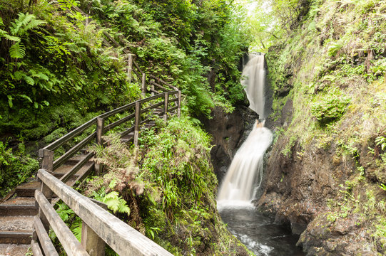 Footpath outdoor trail track with wooden fence next to a small fast flowing waterfall in a gorge in the forest. Glenariff Forest Park, Northern Ireland, UK.