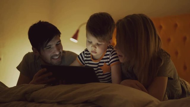 Happy family with little son learning to play tablet computer lying in bed at home in evening