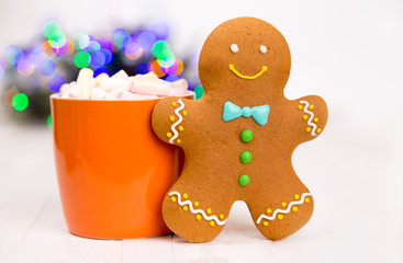 Fototapeta na wymiar Smiling gingerbread man standing next to orange mug and new year light bokeh. Wihte background. Copy space for text.