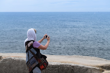 Frau mit Kopftuch fotografiert den Meerblick von der Küste mit dem Handy