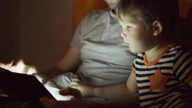Young father and his little son watching cartoon movie using tablet computer and talking while lying in bed in evening at home