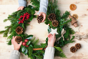 Two small children's hands holding and helping to decorate handmade Christmas wreath made with fir branches, pine cones, berries, baubles on the natural wooden table, top vew, selective focus © Liliya Trott