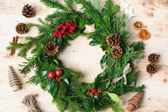 Decorated handmade Christmas wreath made with fir branches, pine cones, berries, baubles on the natural wooden table, top vew, selective focus