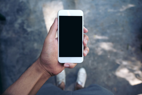 Mockup image of a man's hand holding white mobile phone with blank black screen while standing on concrete polishing floor