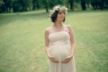 Fototapeta na wymiar Happy pregnant woman in a soft beige dress and a flower wreath on the head walks around the spring park