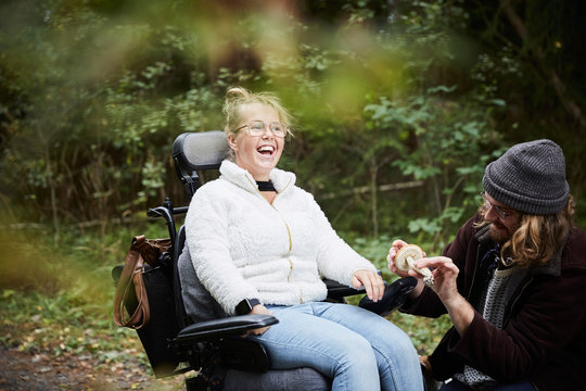 Cheerful disabled woman in wheelchair by caretaker holding mushroom at forest
