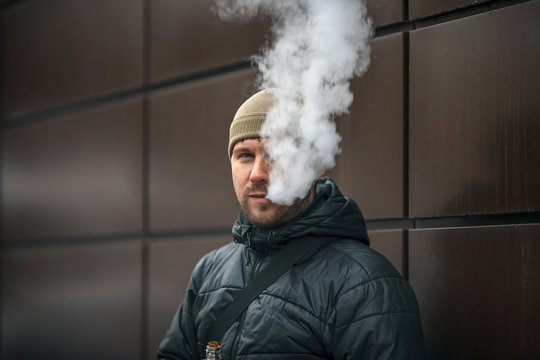 Vape man. Portrait of a handsome young white guy in a modern cap vaping an electronic cigarette opposite the futuristic urban background. Lifestyle.