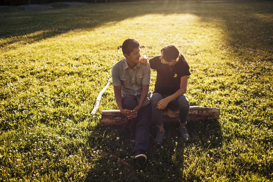 Father and daughter talking while sitting on log amidst grassy field at park during sunny day