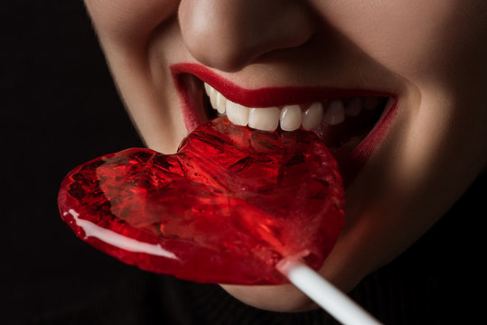 cropped image of woman biting heart shaped lollipop isolated on black