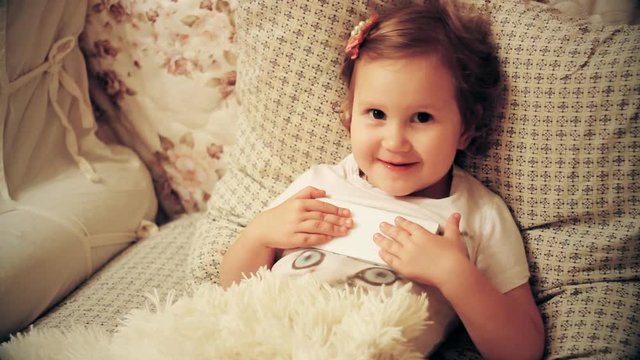 Portrait close-up funny child looks at the phone screen and plays downloaded application on a smartphone close-up. A little cute girl lies in bed in the bedroom under the blanket and playing the game